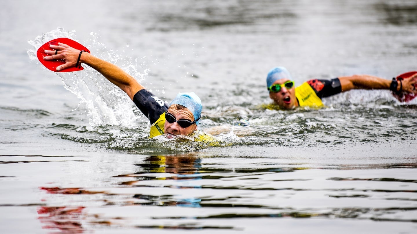 Två swimrunåkare i full sim med röda paddlar, simmössor och tävlingsvästar